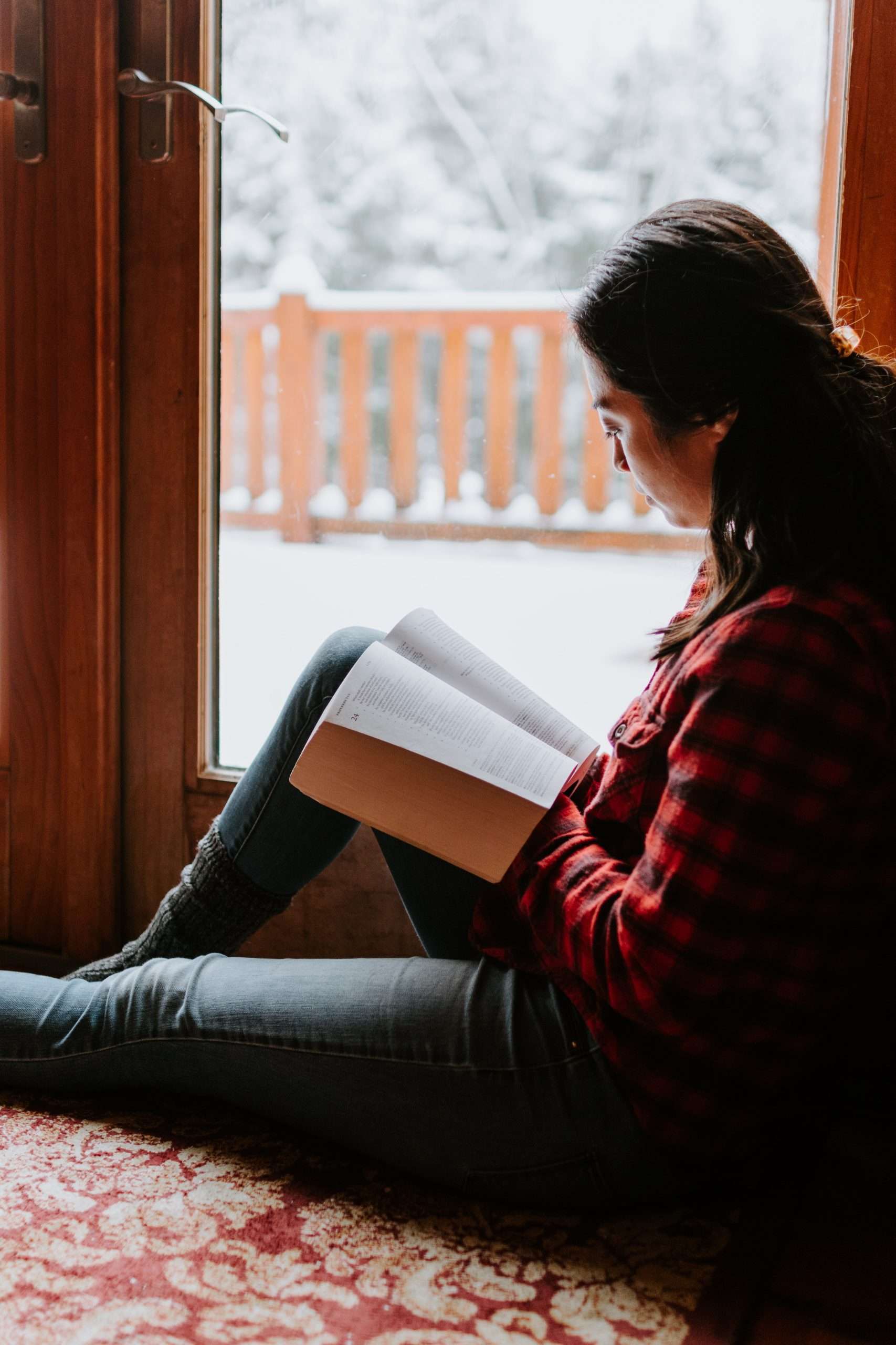 Seated woman reading book near the window with snow outside.